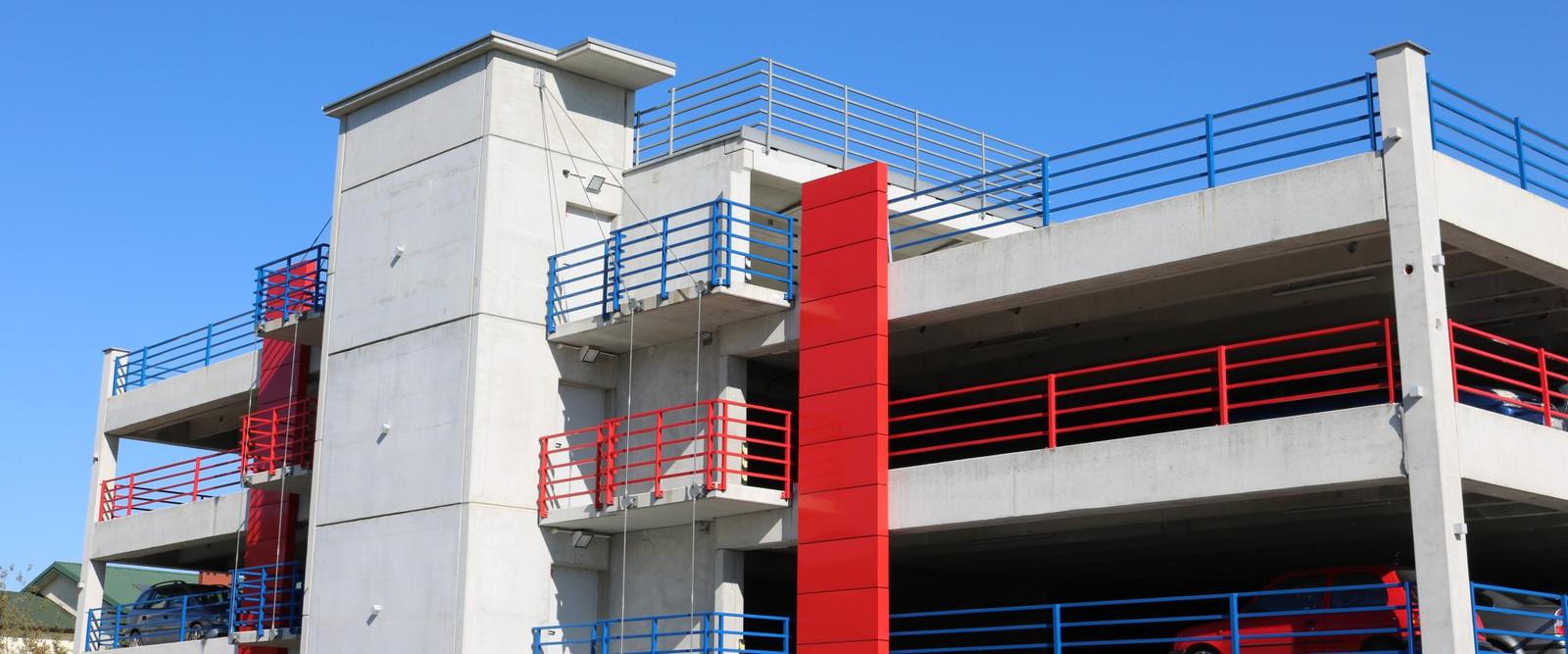 Wielopoziomowy parking modułowy z betonu, z czerwonymi słupami i niebiesko-czerwonymi balustradami.